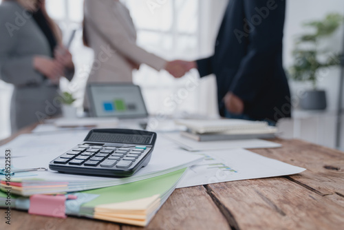 Calculator and documents are lying on a wooden desk in an office, with business people shaking hands in the blurred background, suggesting a successful business deal or partnership