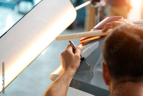 Inside a Garage, an automotive technician is shown from the shoulders up using a specialized tool to repair a dent on a gray automobile. A wooden brace and light are used