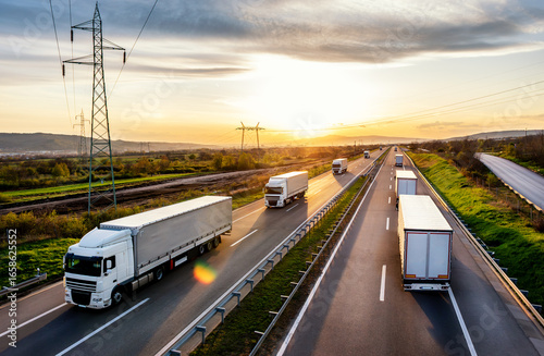 Highway transportation scene with Convoys of white transportation trucks passing each other on a rural highway at a beautiful sunset sky