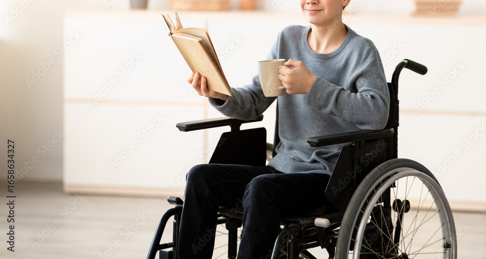 Fototapeta premium Positive impaired teenager in wheelchair drinking tea and reading interesting book at home. Happy handicapped adolescent relaxing, enjoying hot beverage and cool story indoors