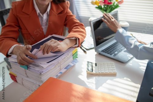 businesswoman and her secretary are helping search for documents examine large number investment documents piled on desk. businesswoman is having trouble finding large number documents piled on desk.