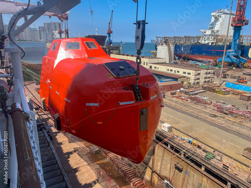 Enclosed orange lifeboat being lowered at a shipyard with cargo ship and cranes in the background.