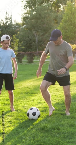 Father and son play soccer on green lawn during sunset. Dad and child are running, smiling, passing ball to each other. Happy family time, outdoor activity, joy, active lifestyle in nature. Vertical.