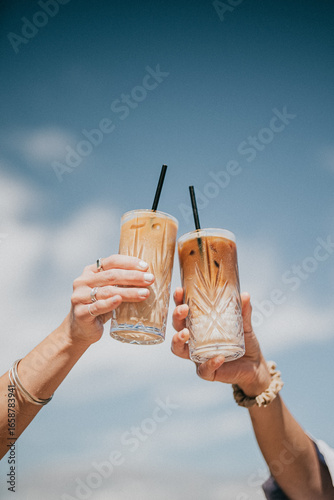 Two people are holding up two glasses of coffee, one of which has a straw in it