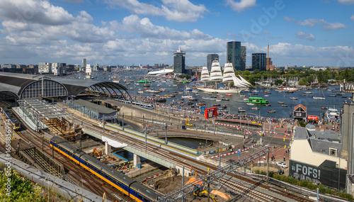 Photography A vibrant view of Amsterdam Central Station with NS trains, trams, and a large crowd gathered along the IJ river during a maritime event featuring numerous boats and a tall sailing ship
