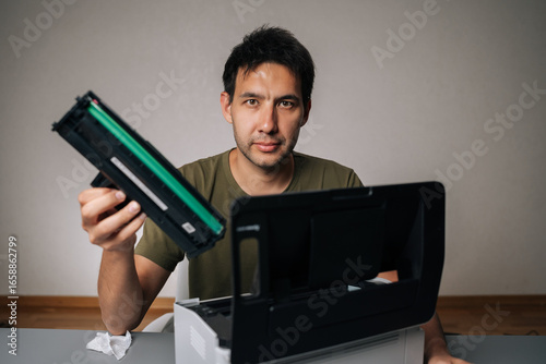 Billede på lærred Studio portrait of computer technician replacing toner cartridge in laser printer during maintenance service, holding new cartridge and looking at camera, sitting at table on isolated background