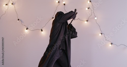 A male actor in a black death suit waves his scythe menacingly in the studio. Halloween concept, black cloak with grim death scythe. Horror robe costume or ghost. Scream costume
