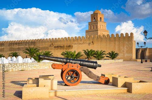 A cannon in front of the city walls of Kairouan, with a cemetery visible in front of them and the Great Mosque also known as the Mosque of Uqba in the background, Tunisia.