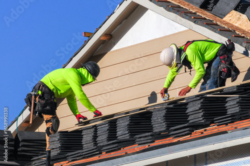 Roofers install rows of stacked shingles on a new house under construction, wearing safety gear and bright protective clothing as part of the residential building process