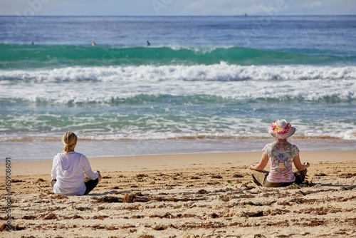 Two ladies, with their backs to the viewer, sit in yoga poses looking toward the ocean waves.