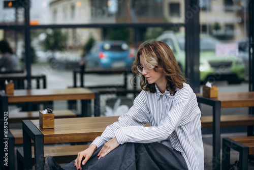 A teenage girl model is young and beautiful in a shirt and trousers on the street in a street cafe