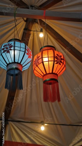 Two illuminated paper lanterns, one blue and one orange, hang from a wooden structure with decorative flower cutouts and tassels.
