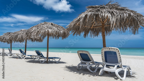 Photography A row of plastic deck chairs under straw umbrellas on a sandy beach