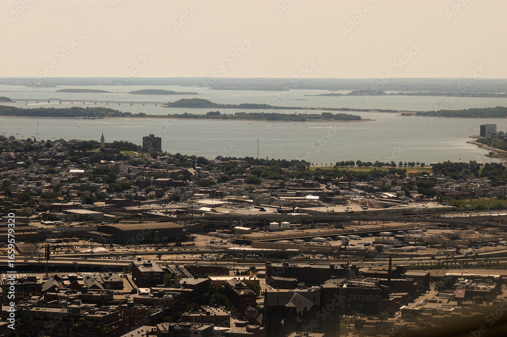 Fototapeta premium Dive into this vibrant aerial view of a coastal urban landscape. The city boasts a grid-like layout of residential and industrial buildings, split by a river or bay. A prominent bridge stretches