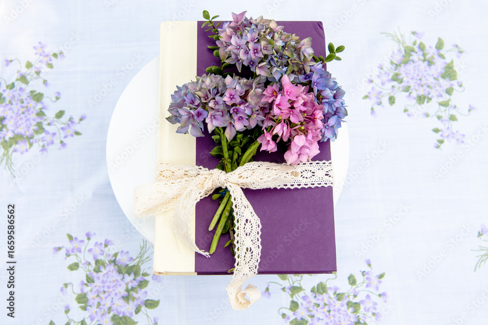 Obraz premium Charming floral book centerpiece with hydrangeas and lace ribbon, set against a lush backyard backdrop for garden-themed events and literary gatherings.