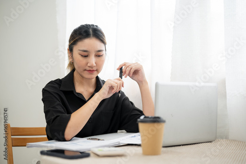 Serious asian businesswoman office worker holding pen reading working document with laptop on table.