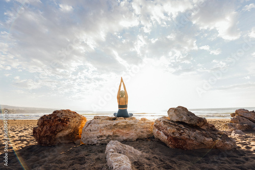 Woman practicing yoga sitting on rock at beach in front of sky