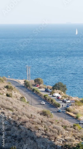 Wallpaper Mural Vertical shot from high angle shows coastal road, dry hillside, parked cars, streetlights and calm blue sea with sailboat and ship on horizon in Vouliagmeni, Athens, Greece, summer daylight Torontodigital.ca