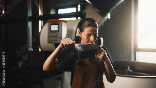 Fotografi Caucasian woman in brown apron holding metal scoop close to face and smelling freshly roasted coffee beans