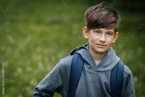 Boy in a gray hoodie with a blue backpack stands in a grassy field, posing with one hand on his hip and looking confidently at the camera.