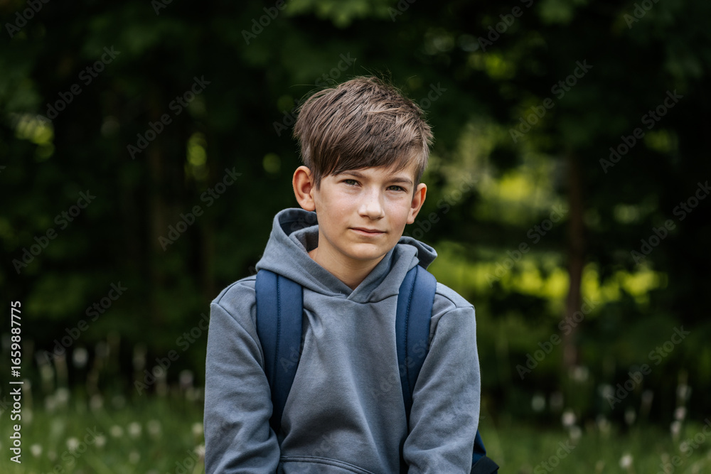 Close-up portrait of a boy in a gray hoodie with a blue backpack, looking calmly at the camera with a natural green background outdoors. Close-up portrait of a boy in a gray hoodie with a blue backpack, looking calmly at the camera with a natural green background outdoors.
