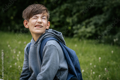Smiling boy in a gray hoodie with a blue backpack squints playfully outdoors, sitting on grass with greenery in the background.