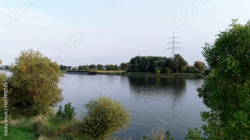 Drone flight at low altitude over a green meadow on the banks of a wide river at sunset. The light highlights the natural structure of the landscape and creates a calm and peaceful view of the scenery