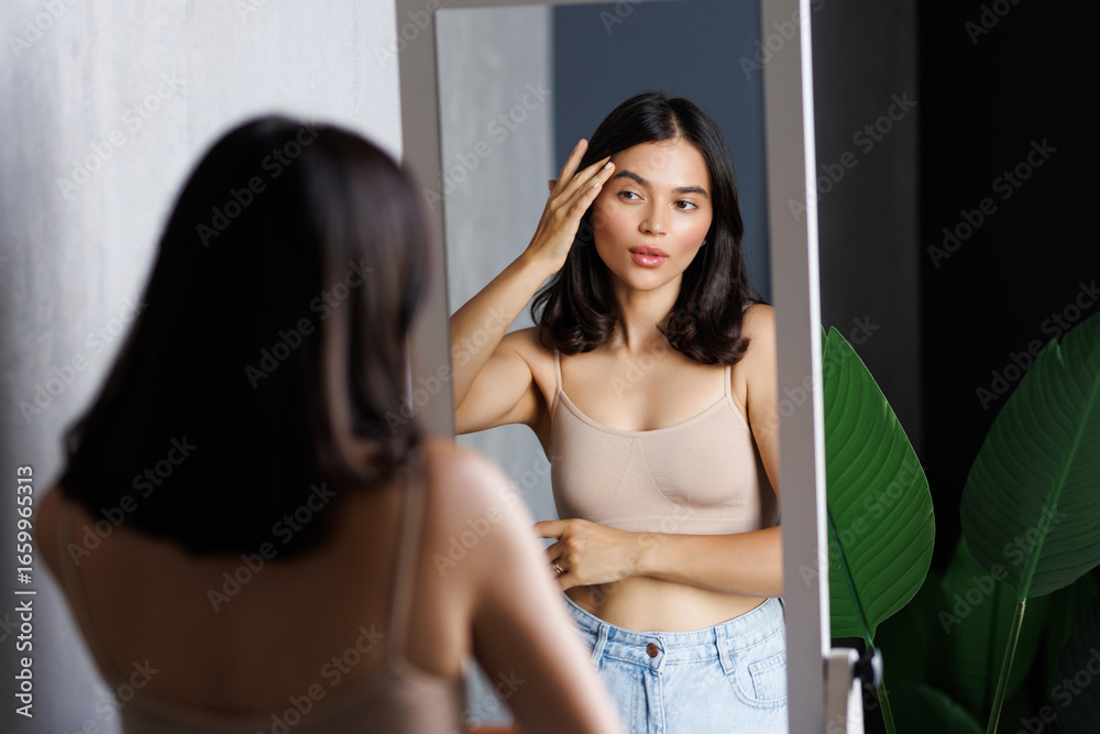 Fototapeta premium Happy beautiful young lady touching face look in bathroom mirror