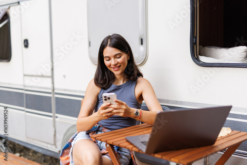 Young woman behind laptop with phone working near trailer.