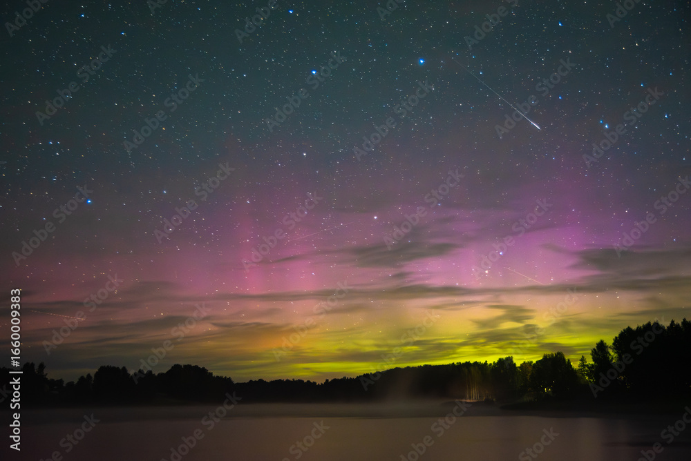 Fototapeta premium Colorful aurora borealis in green and pink shades over a calm lake with forest silhouettes and a shooting star in the starry night sky.