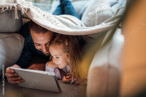 Happy man sharing tablet computer with daughter under blanket at home