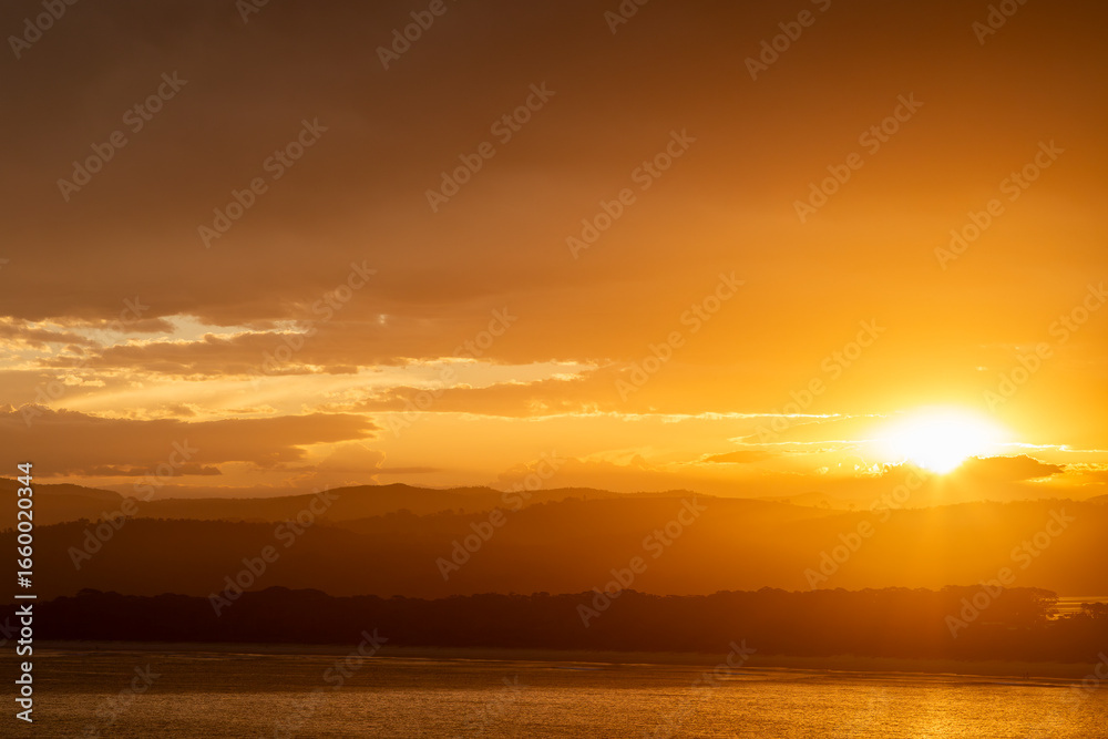 Obraz premium Fiery sunset at Merimbula, Sapphire Coast New South Wales, Australia. Orange sky glowing beneath clouds across coastal hills, calm ocean and lake, creating a dramatic, tranquil seascape at dusk.
