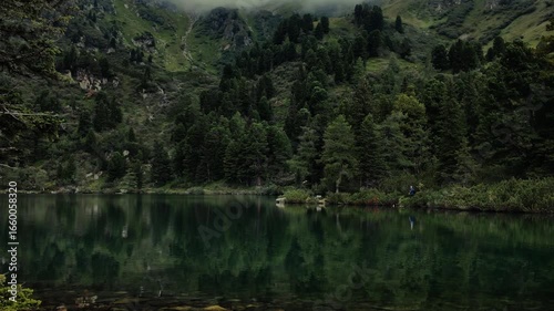 A stunning, amazing view of a mountain lake on a rainy and foggy day. The deep green surface perfectly reflects the surrounding forest and alpine peaks, shrouded in clouds.