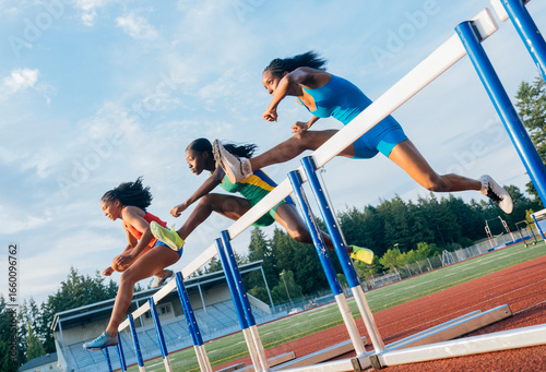 Three athletes jumping hurdles on a track under a clear blue sky during a race. USA