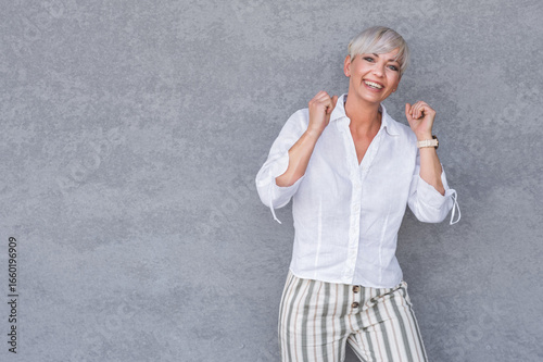 Amused beautiful adult woman with closed eyes and clenched fists, against gray background - wall. Yes concept. Amusing middle age woman celebrates success