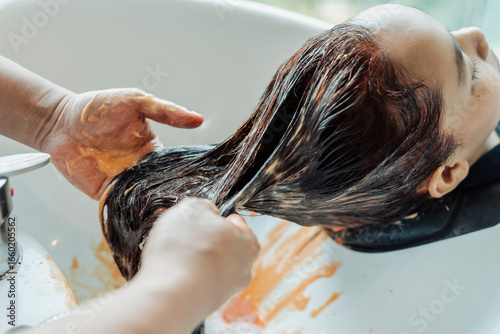 Detailed view of Southeast Asian woman’s hair as a stylist rinses out red hair dye after coloring at a beauty salon.