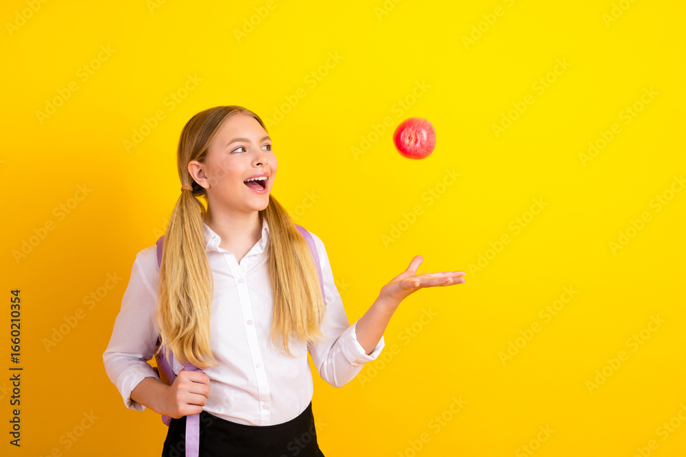 Cheerful schoolgirl with backpacks and pigtails joyfully holding a red apple against a vibrant yellow background Cheerful schoolgirl with backpacks and pigtails joyfully holding a red apple against a vibrant yellow background