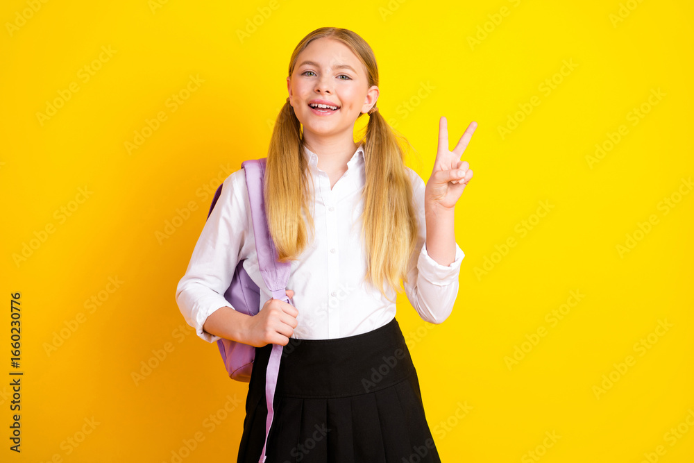 Smiling young schoolgirl with pigtails holding backpack and showing a peace sign on vibrant yellow background Smiling young schoolgirl with pigtails holding backpack and showing a peace sign on vibrant yellow background