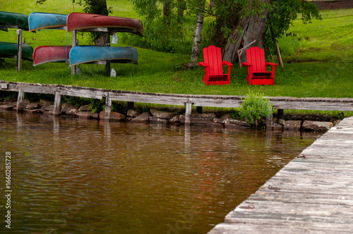 Two red Adirondack chairs and six canoes upside down on a rack by the shoreline of lake. the water and a dock are in the foreground with grass and trees in the background during the summer.