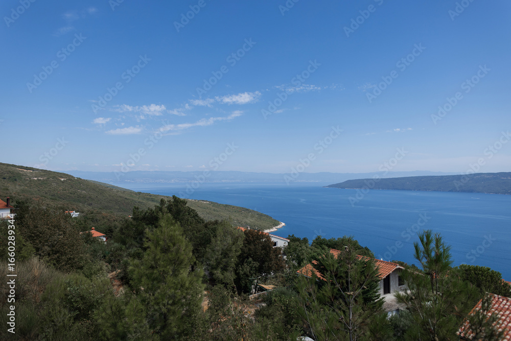 Fototapeta premium Scenic view of the Adriatic Sea with red-roof houses in a coastal Croatian village under a clear blue sky. Peaceful seascape with gentle waves and wide horizon under a clear sky in Croatia.