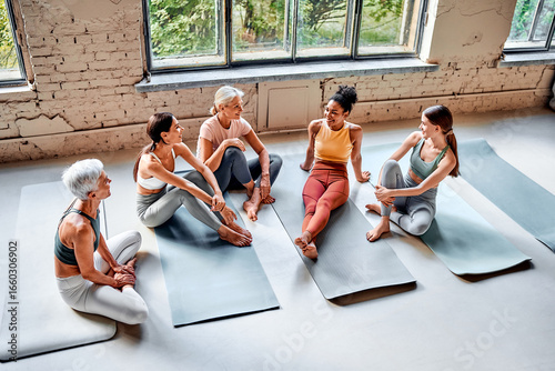 Diverse women are sitting on mats in the studio, relaxing and chatting after a workout. Friends at the fitness studio.