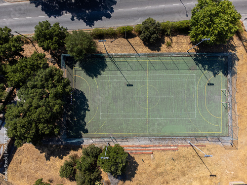Aerial view of an outdoor futsal court in a playground, surrounded by green trees and open space, captured on a sunny day.