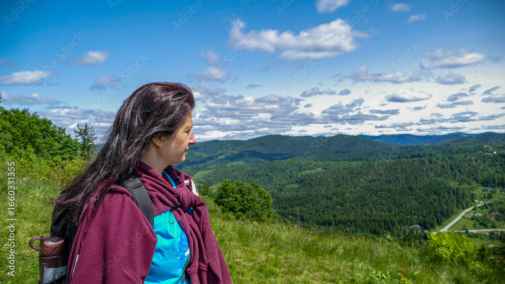 Naklejka premium woman 40 years old, with a brown backpack, dark hair, looking at mountain, tourists, beautiful landscape, hike, protected area, sky, clouds, forest, various grasses, space, summer, hike,