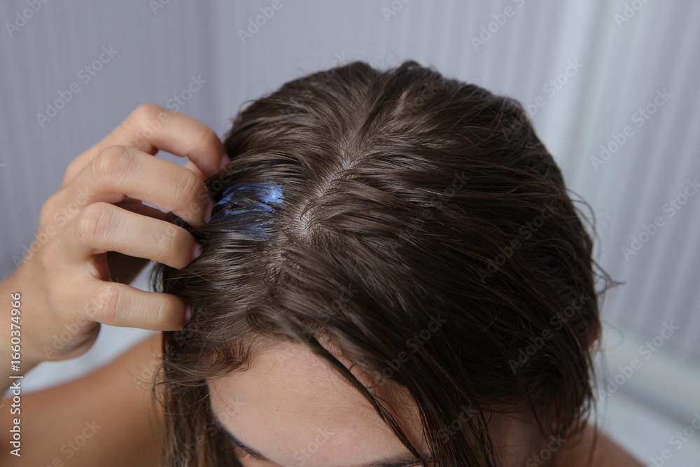 Fototapeta premium A person is shown applying a blue product directly to their wet, brown hair with their fingers, with a blurred bathroom background