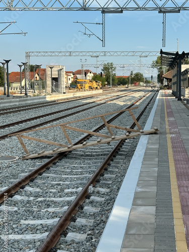 A newly constructed railway station platform is shown with train tracks, overhead electrical structures, and a wooden barrier placed across the rails