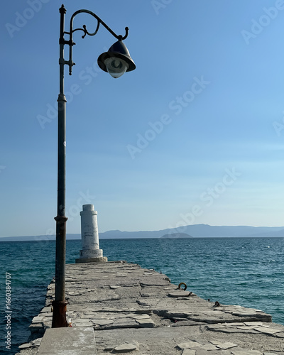 A rustic stone pier with an old street lamp and a small white lighthouse extends into the blue sea under a clear sky