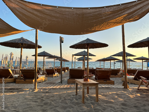 A serene beach scene with wooden sunbeds, straw parasols, and a canopy shade at sunset by the sea
