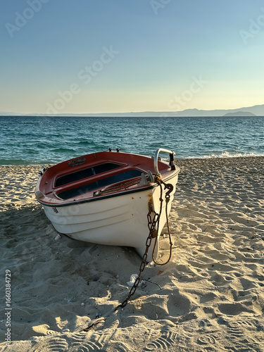 A small white and red boat with a chain is resting on the sandy beach by the sea at sunset