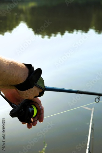 A close-up shot of a person holding a fishing rod, with a visible fishing reel and bright green fishing line. The photo is taken outdoors by a peaceful body of water with trees in the background.