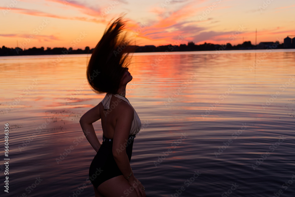 Fototapeta premium Woman in black bikini stands on shoreline at sunset, hair whipping in wind. She poses with hand on hip, facing vibrant orange and pink sky over calm lake.